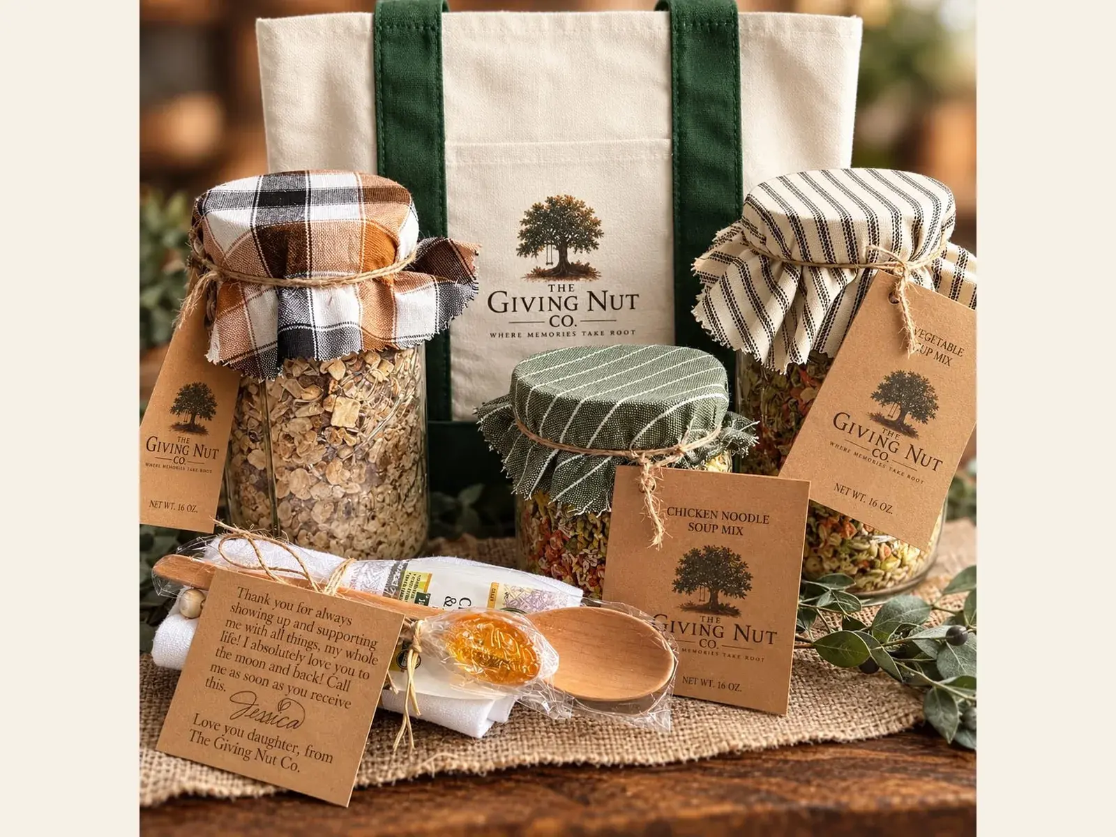 Three Giving Nut Co. jars of soup mix with fabric tops and kraft tags, a canvas tote bag, a wooden spoon and a handwritten gift note, arranged on a burlap runner