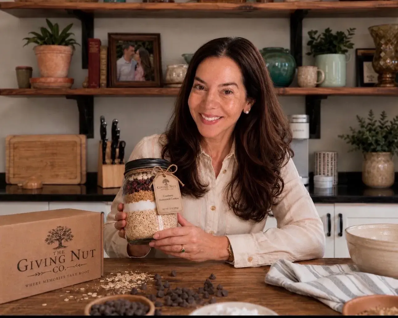 Jessica in her kitchen, smiling, holding a Giving Nut Co. jar of layered cranberry honeycrisp cookie mix, with a branded kraft gift box on the counter beside her